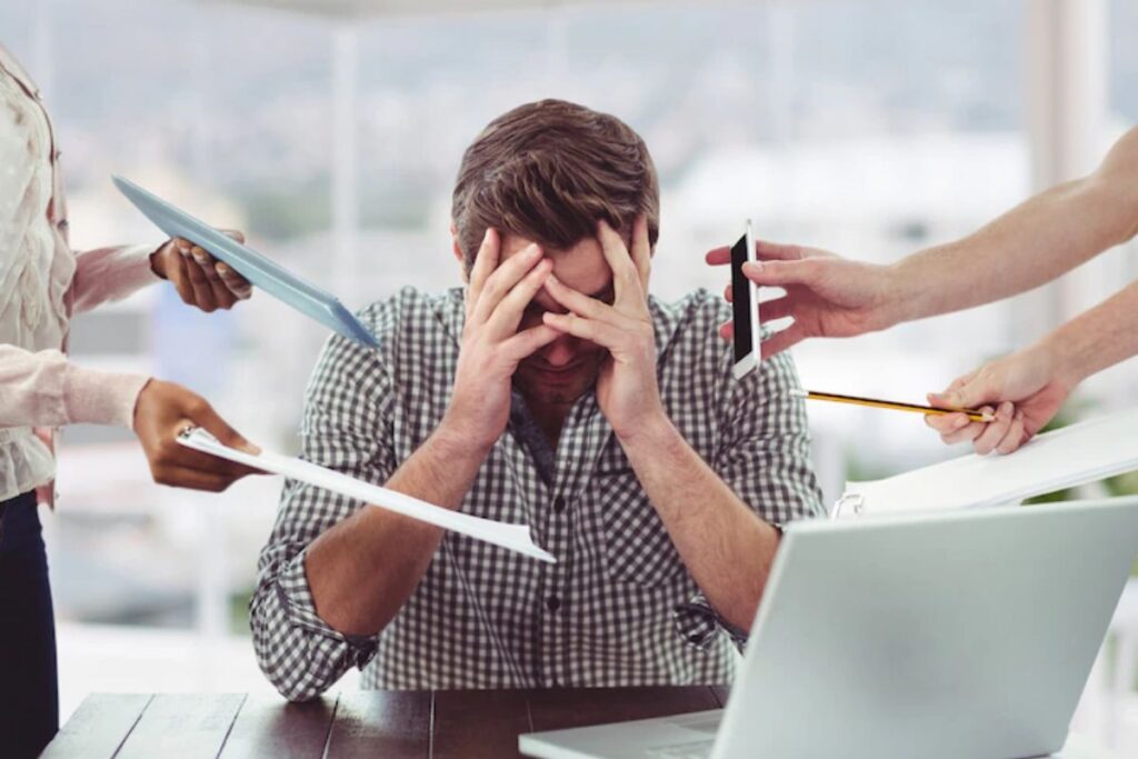 Homem em empresa sentado diante de notebook com pessoas entregando objetos para ele. O mesmo leva as mãos a cabeça como sinal de esgotamento e alerta de falta de saúde mental no trabalho.