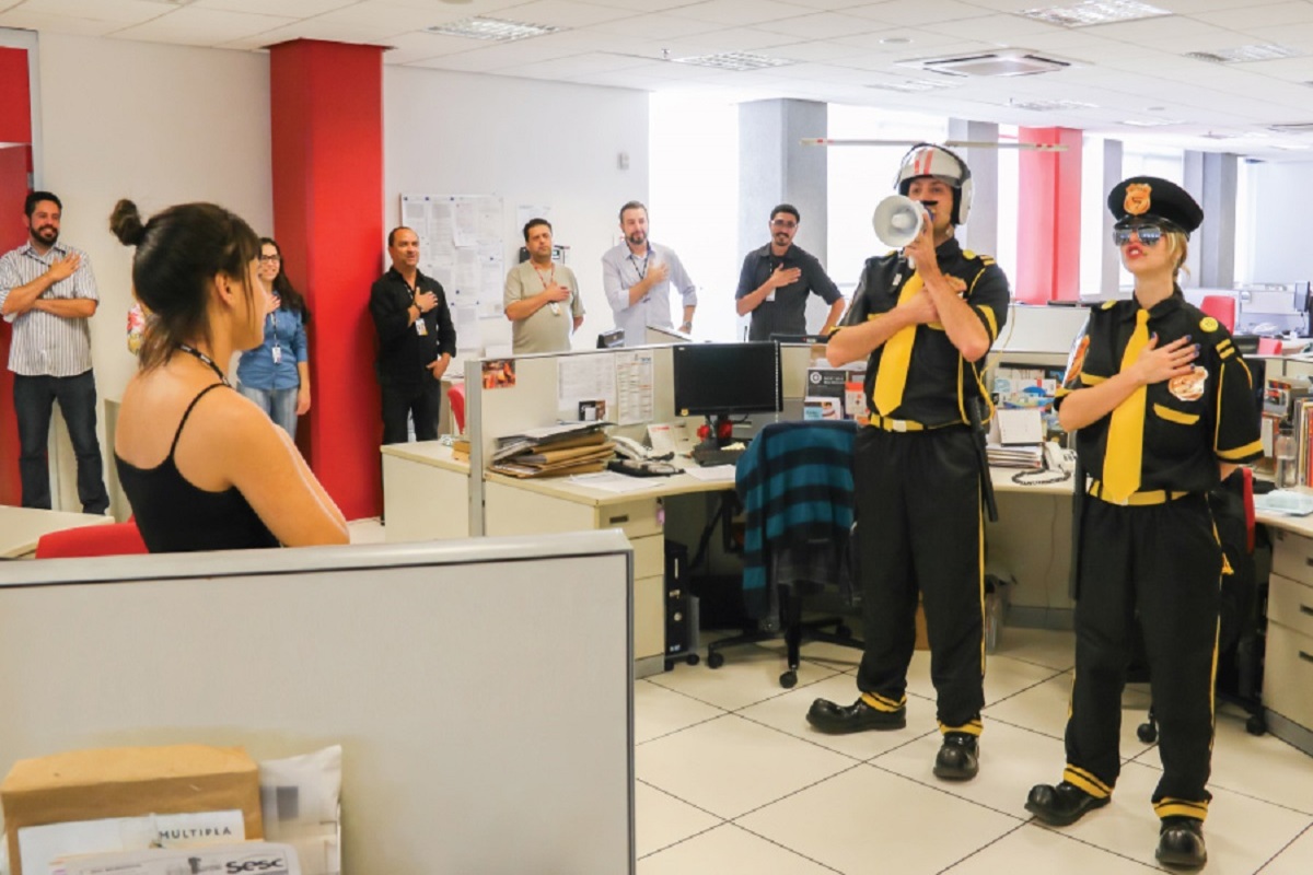Imagem de funcionários em uma palestra com comediantes, todos com a mão no peito como se estivessem fazendo um juramento