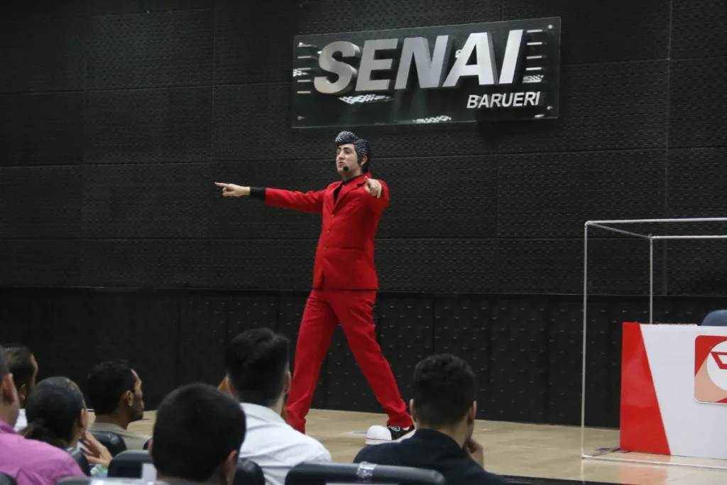 Palestrante vestido de vermelho durante evento da SIPAT no SENAI Barueri falando para colaboradores em auditório.