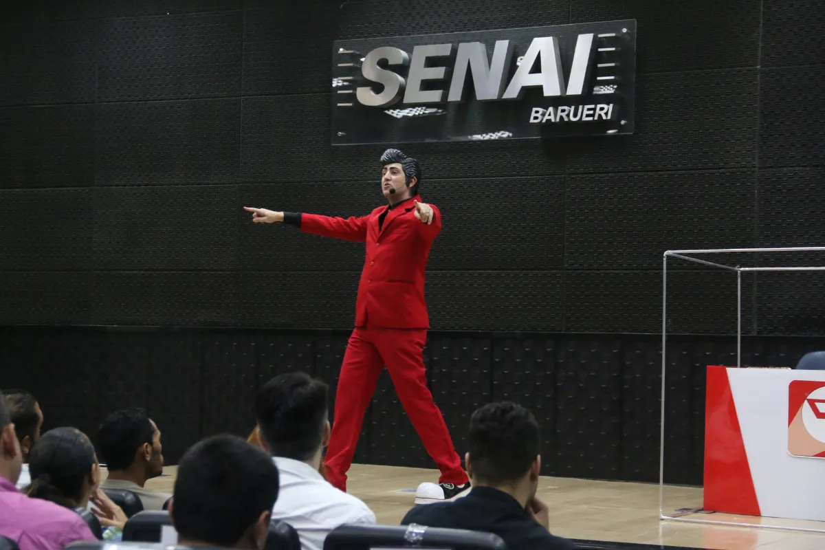Palestrante vestido de vermelho durante evento da SIPAT no SENAI Barueri falando para colaboradores em auditório.