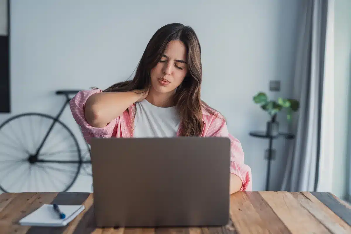 Mulher sentada de frente para um laptop, com uma das mãos na nuca