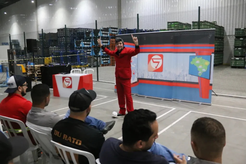 Apresentador conduz dinâmica para colaboradores durante evento de segurança do trabalho dentro de um galpão industrial.