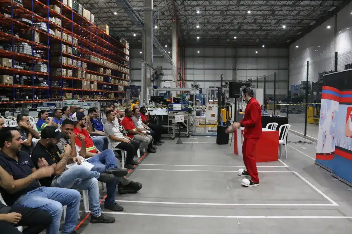 Trabalhadores sentados assistindo apresentação durante evento corporativo em galpão industrial.