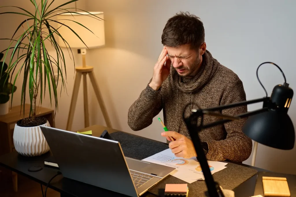 Homem sentado à mesa de trabalho com laptop, segurando a cabeça enquanto analisa documentos
