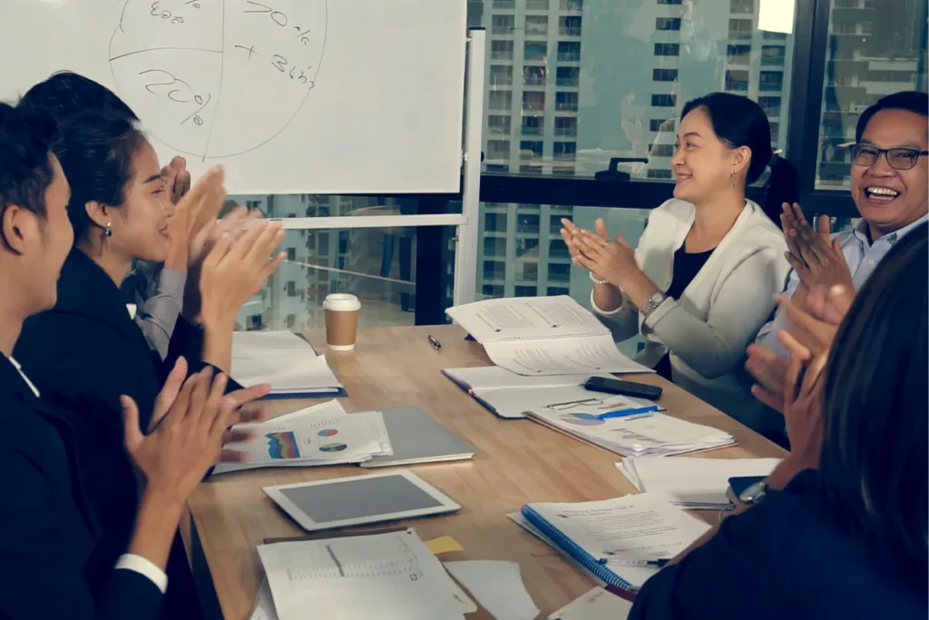 Equipe de colaboradores reunida em sala de reunião aplaudindo durante atividade de treinamento.