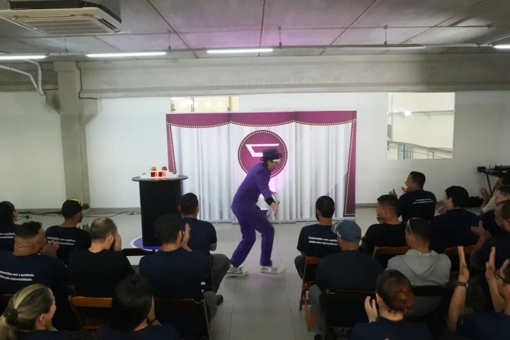 Apresentador vestido de roxo realizando apresentação interativa diante de colaboradores sentados em um auditório interno da empresa.