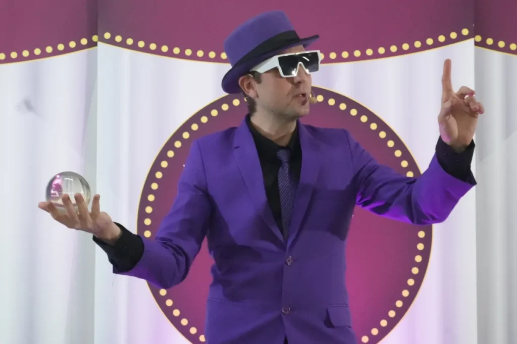 Apresentador usando terno roxo e chapéu durante uma apresentação em palco, segurando uma esfera de vidro e gesticulando para o público.