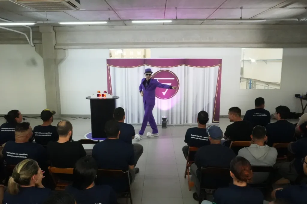 Apresentador vestido de roxo realizando apresentação interativa diante de colaboradores sentados em um evento interno da empresa.