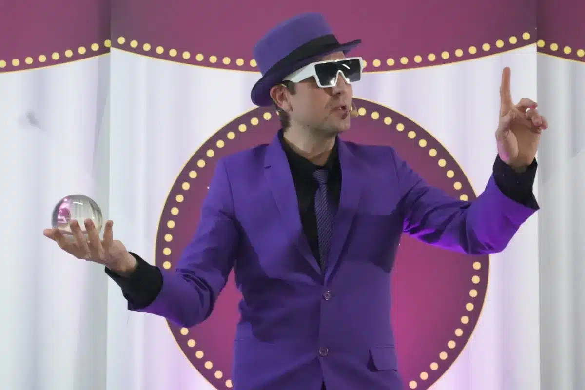 Apresentador usando terno roxo e chapéu durante uma apresentação em palco, segurando uma esfera de vidro e gesticulando para o público.