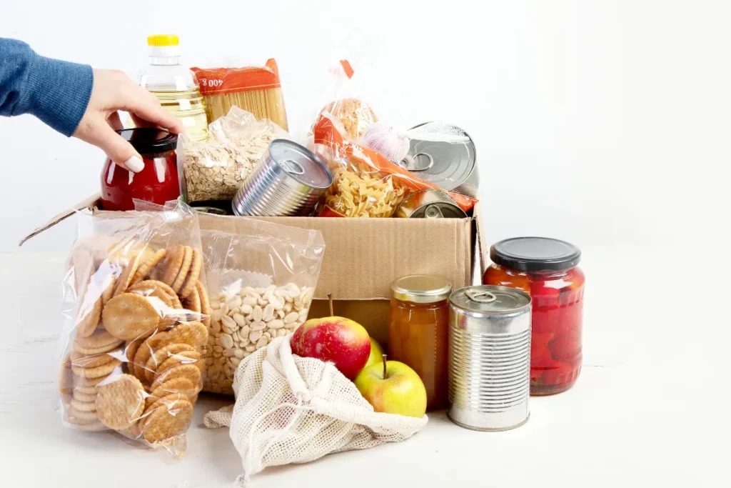 Caixa de papelão com alimentos como enlatados, macarrão e frutas sendo organizada por uma pessoa