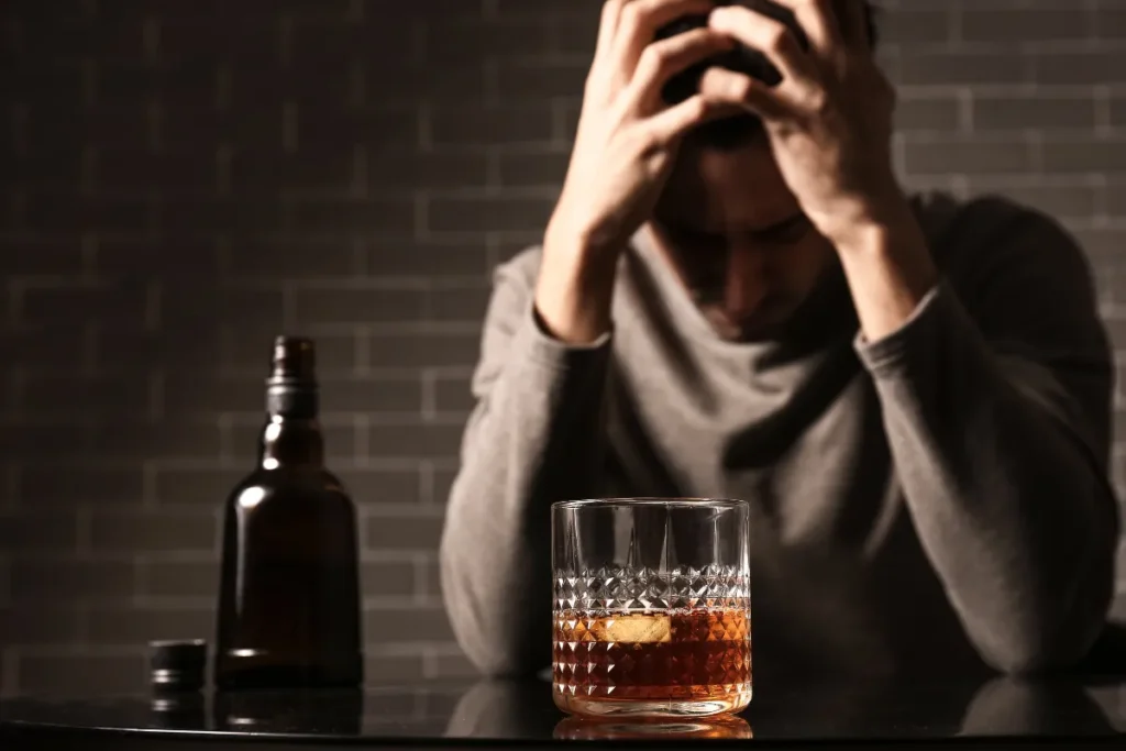 Homem sentado com a cabeça nas mãos ao lado de copo e garrafa de bebida alcoólica