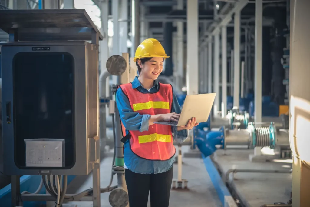 profissional com capacete e colete usando notebook dentro de instalação industrial com máquinas ao fundo