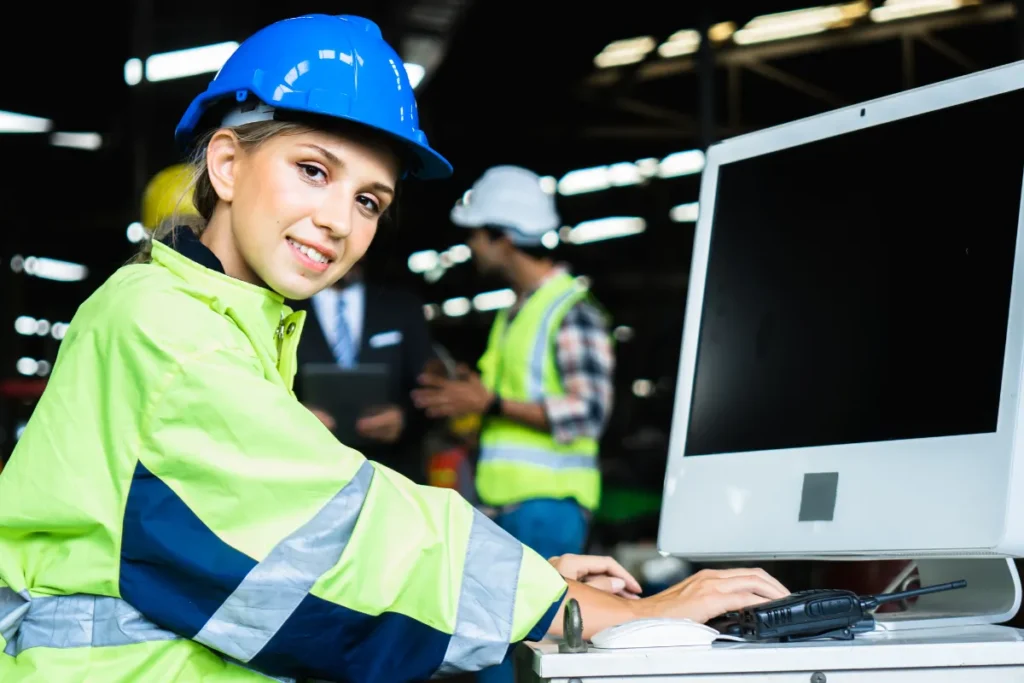 Mulher com capacete azul e uniforme de segurança utilizando computador em ambiente industrial