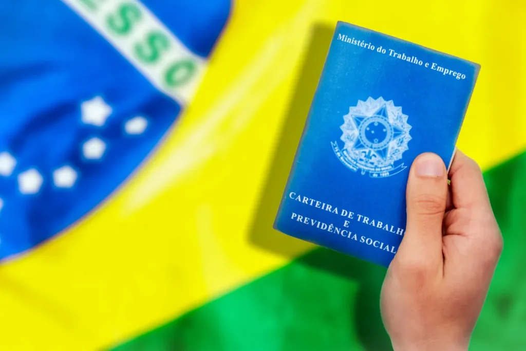 mão segurando carteira de trabalho azul com bandeira do Brasil ao fundo