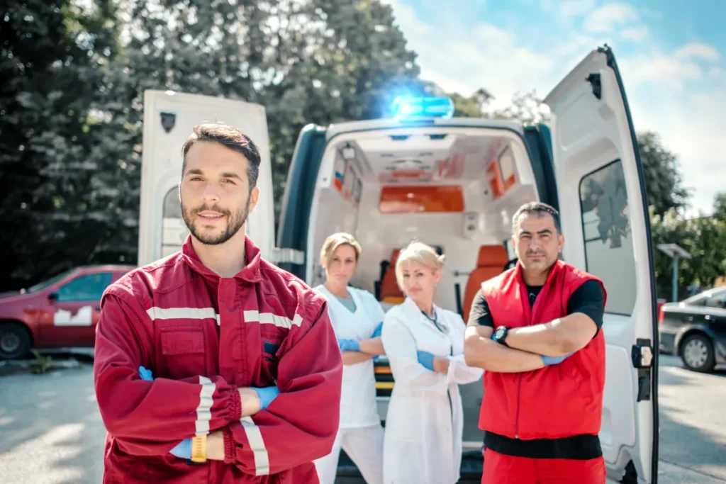 Equipe de emergência com uniformes vermelhos e brancos em frente a ambulância com portas abertas