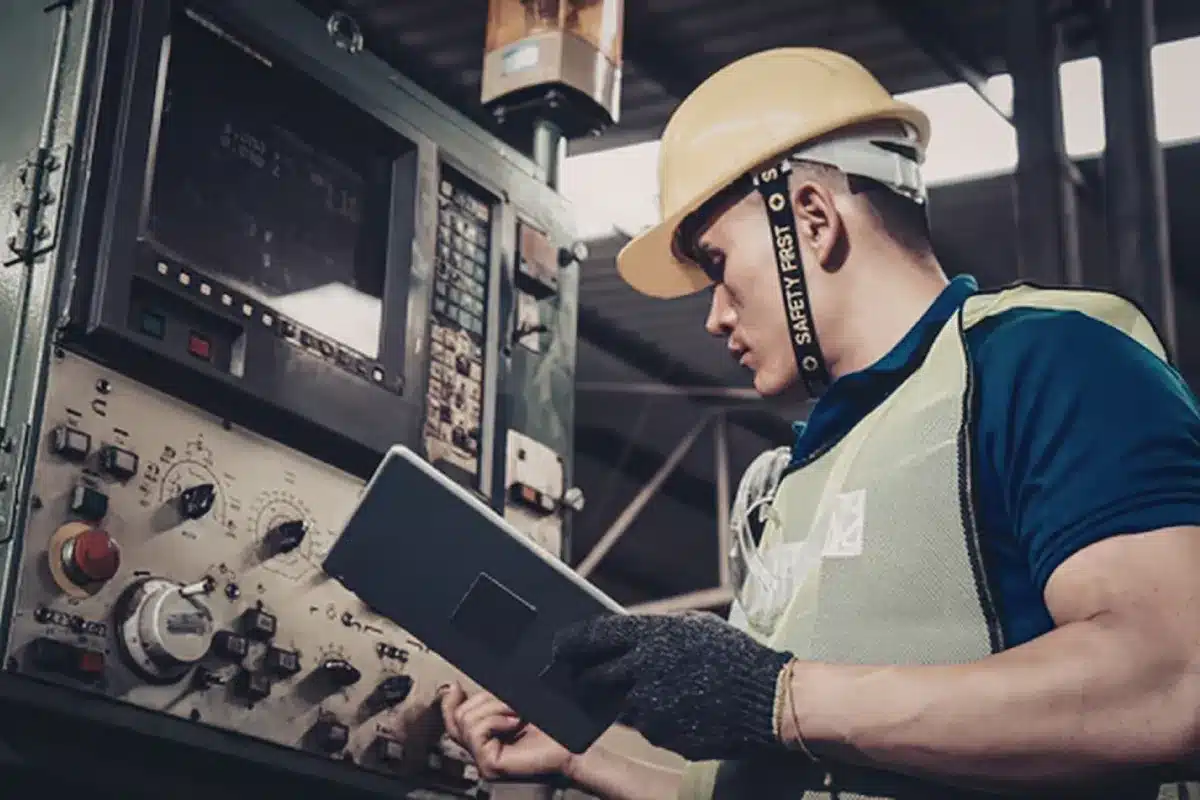 Trabalhador usando capacete e luva opera painel industrial enquanto segura um tablet para monitoramento de equipamentos