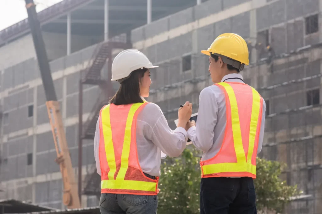 Dois trabalhadores com capacetes e coletes refletivos conversando em frente a obra em construção