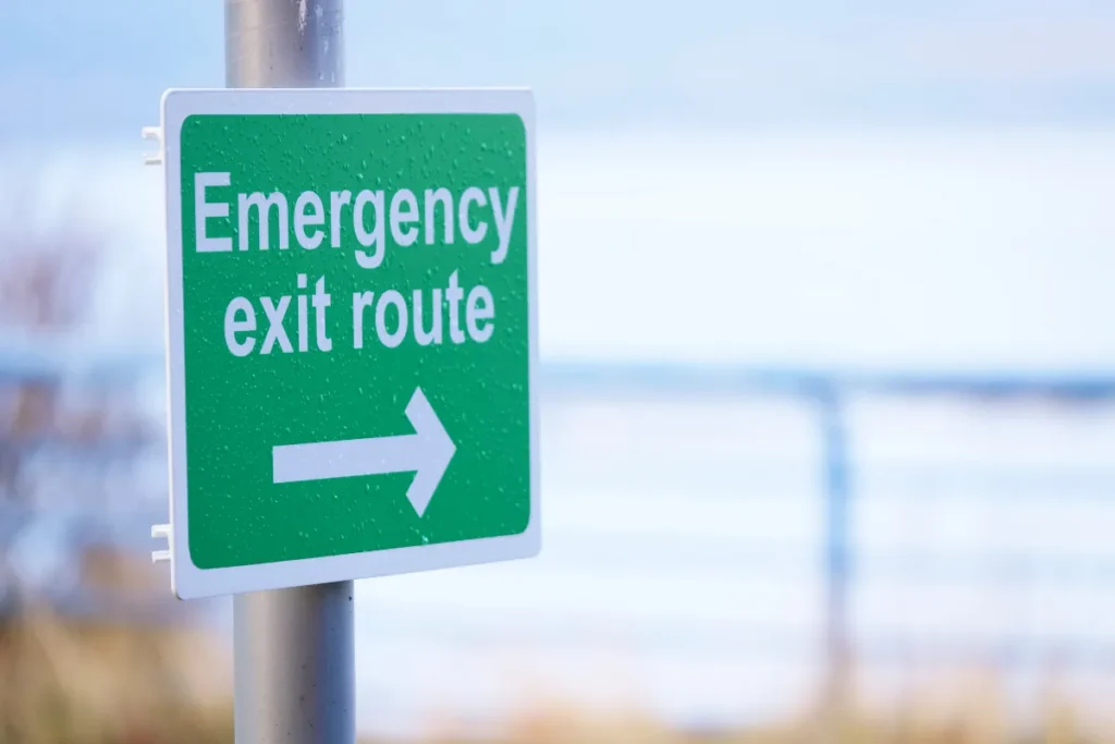 Placa verde de emergency exit route com seta para direita instalada em área externa indicando direção de saída segura em caso de emergência