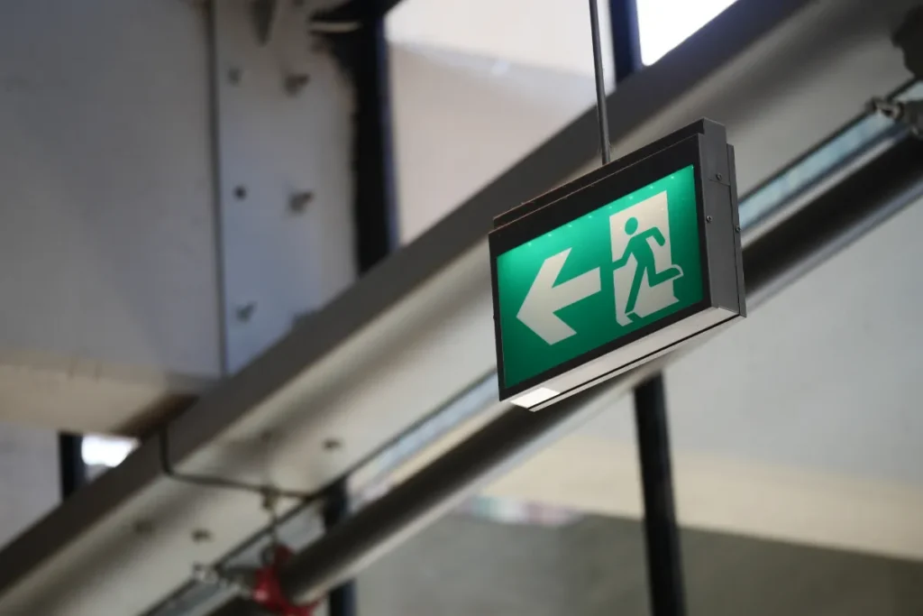 Placa suspensa de rota de fuga com seta apontando para esquerda instalada em estrutura interna com iluminação indicando caminho de evacuação