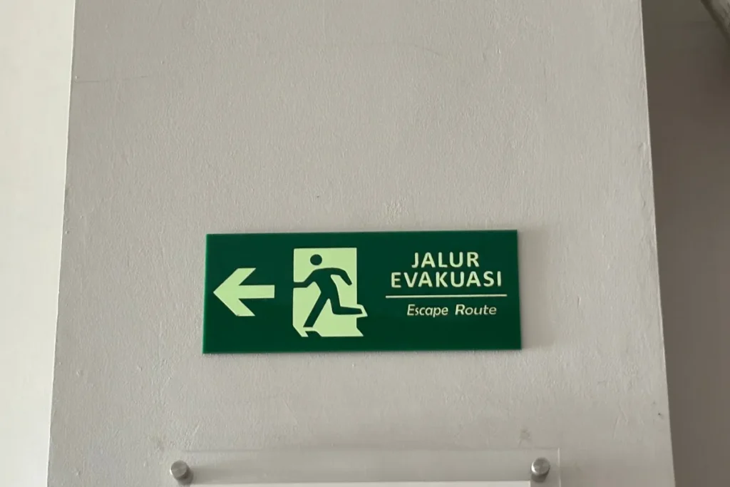 Placa verde de rota de fuga com seta para esquerda fixada em parede branca indicando direção de evacuação em ambiente interno corporativo