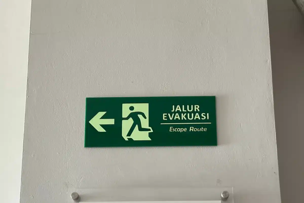 Placa verde de rota de fuga com seta para esquerda fixada em parede branca indicando direção de evacuação em ambiente interno corporativo