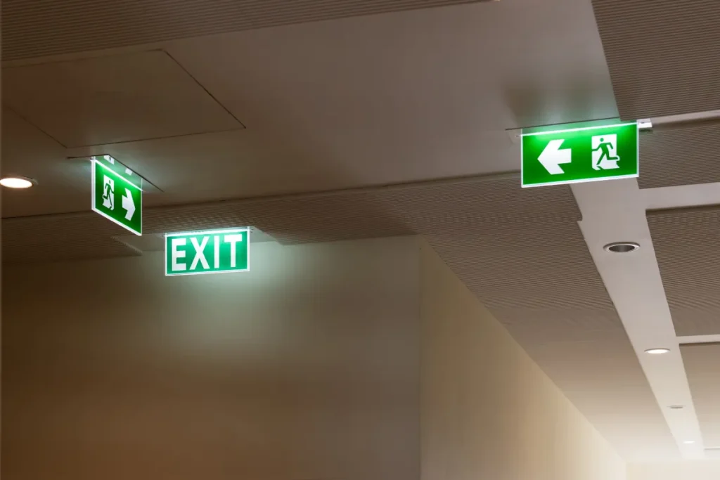 Corredor corporativo com placas iluminadas de saída de emergência e sinalização de rota de fuga no teto indicando direções de evacuação