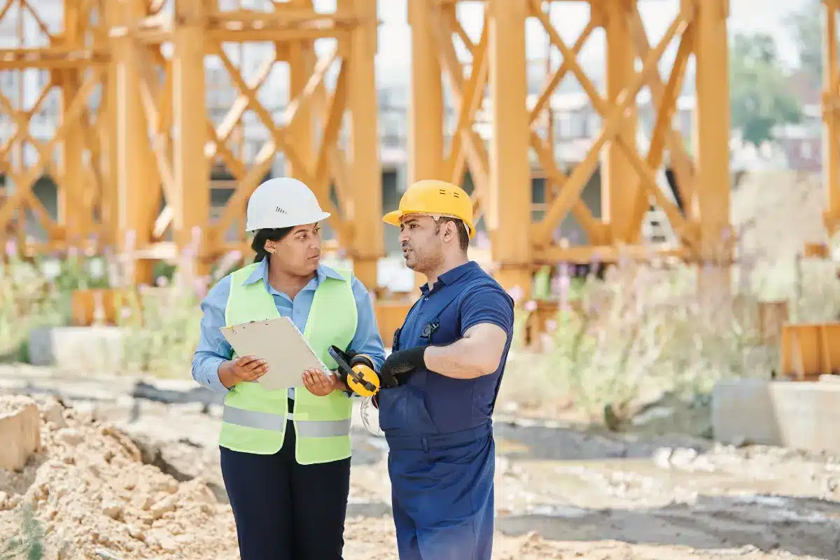 Dois profissionais com capacetes e equipamentos de segurança discutindo informações em um canteiro de obras