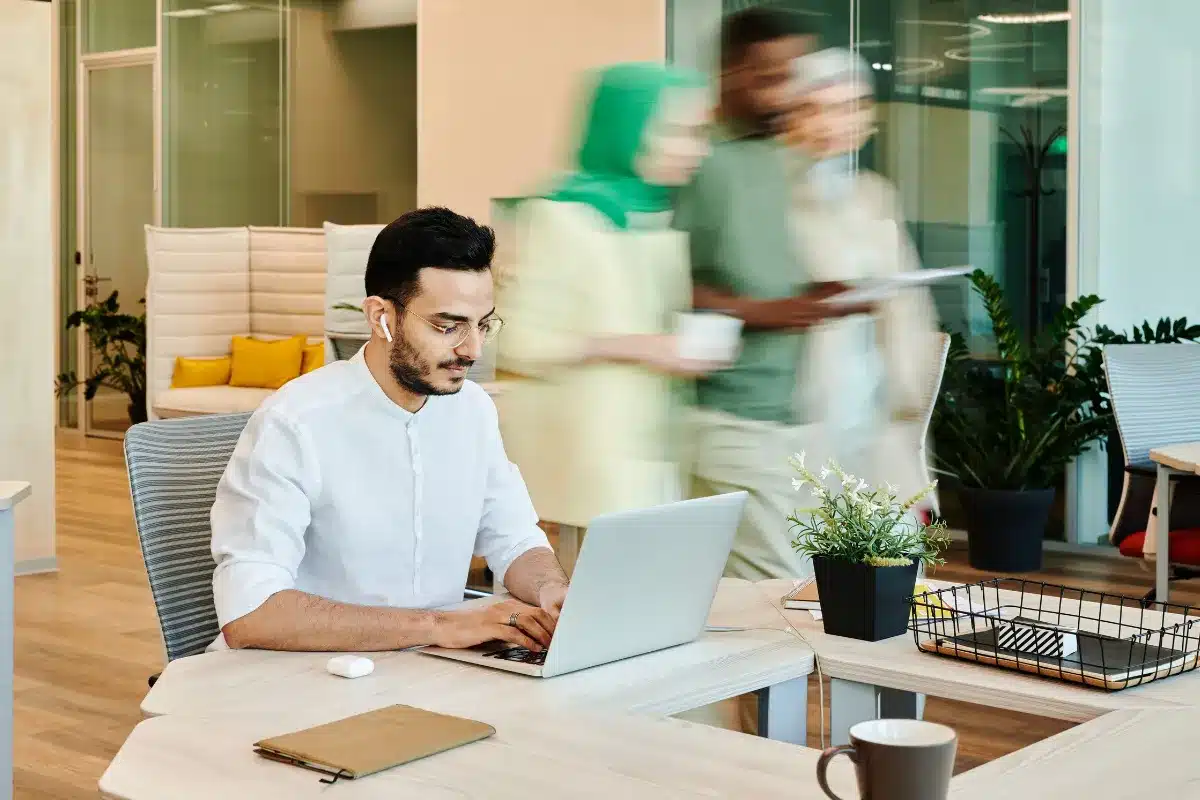 Profissional trabalhando concentrado em um laptop enquanto colegas aparecem em movimento ao fundo