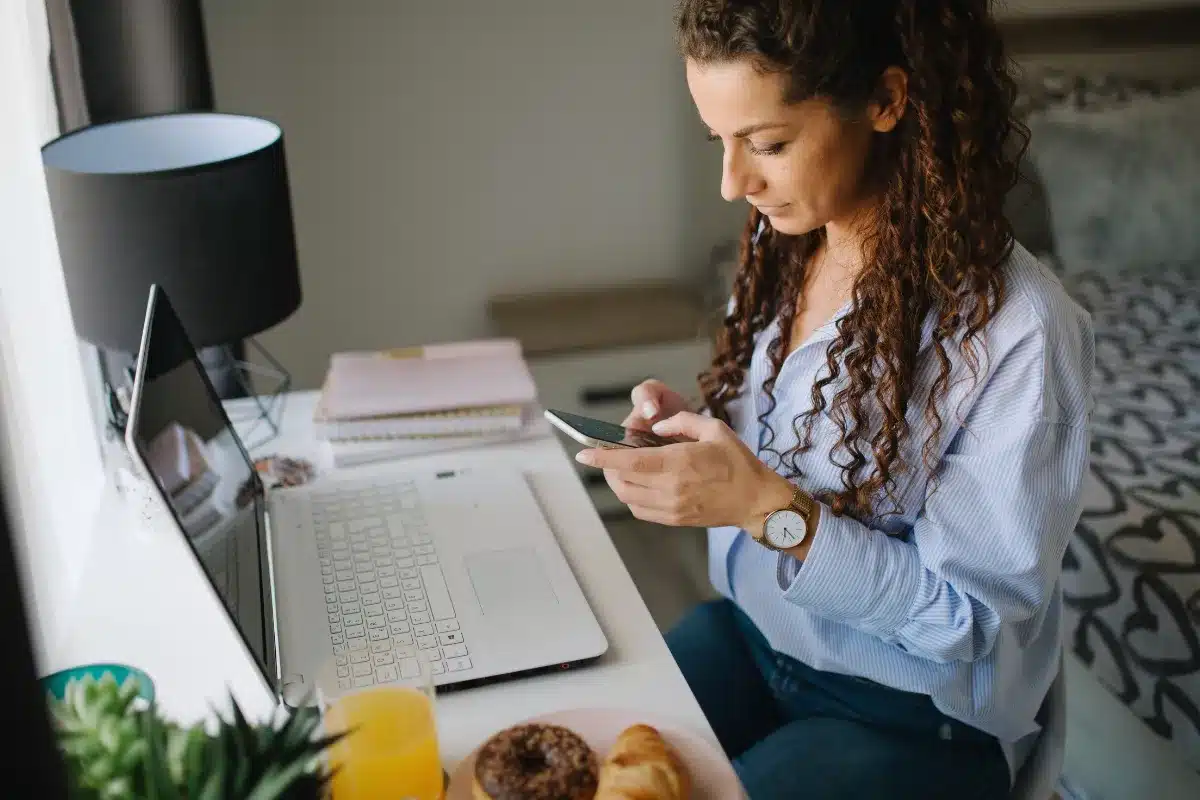 Mulher usando smartphone ao lado de um notebook em uma mesa de trabalho