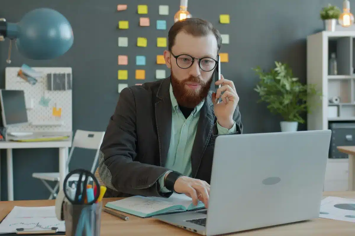 Profissional trabalha no notebook enquanto fala ao celular, ilustrando multitarefa e interrupções frequentes.