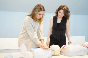 Duas mulheres praticam reanimação em bonecos de treinamento numa sala de curso