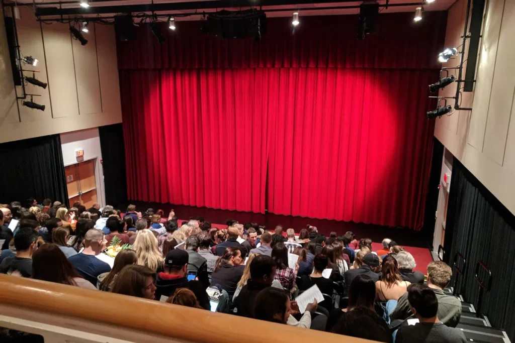 Plateia acomodada num auditório diante de um palco com cortinas vermelhas fechadas.