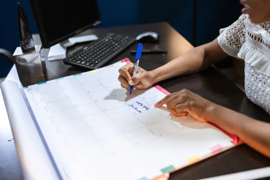 Profissional anota um compromisso no calendário de mesa, ao lado do teclado do computador.