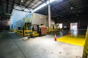 Galpão de armazém com empilhadeira carregando sacos, cones de sinalização e faixa amarela de segurança no piso