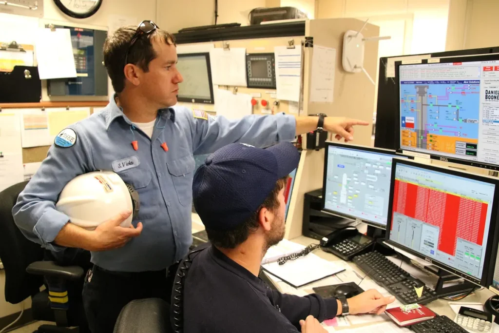Inspetor de segurança com capacete na mão aponta para telas de monitoramento ao lado de um operador