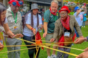 Grupo de adultos ao ar livre segurando varetas juntos para manter uma bola equilibrada durante uma atividade em equipe.