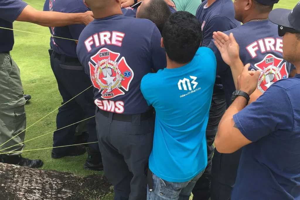Bombeiros e um colega de camiseta azul seguram cordas juntos num exercício de equipe ao ar livre.
