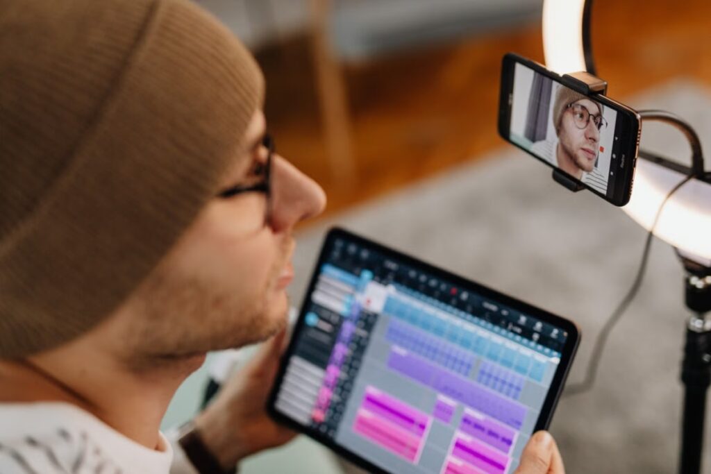 Vista em ângulo superior e lateral de um homem de touca e óculos olhando para um tablet com gráficos coloridos na mão, enquanto é filmado por um smartphone acoplado a um ring light.