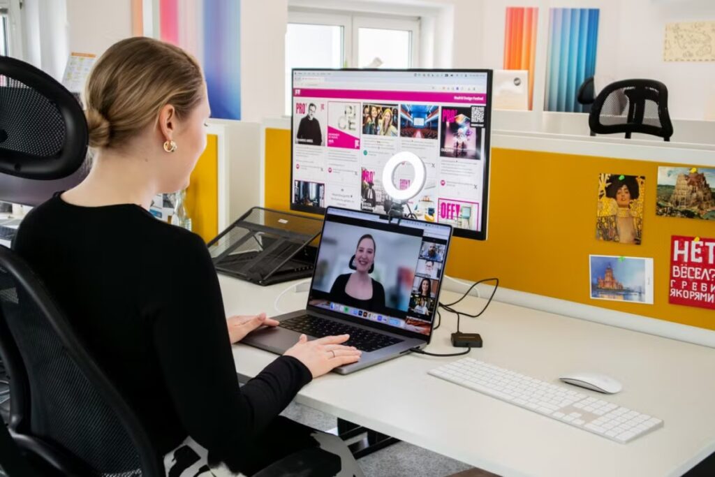 Vista oblíqua traseira de uma mulher loira sentada em uma cadeira de escritório ergonômica preta, assistindo a uma videoconferência em um notebook integrado a um monitor maior com ring light na mesa.