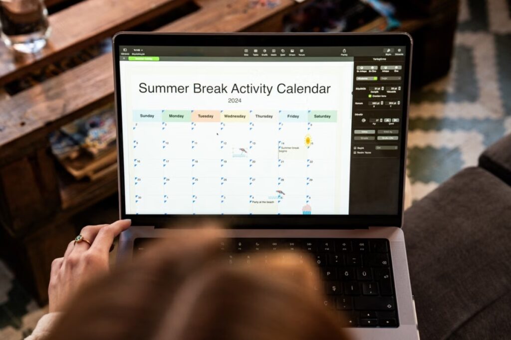 Vista superior em primeira pessoa da cabeça e das mãos de uma pessoa olhando e editando um calendário digital intitulado "Summer Break Activity Calendar" na tela de um notebook aberto.