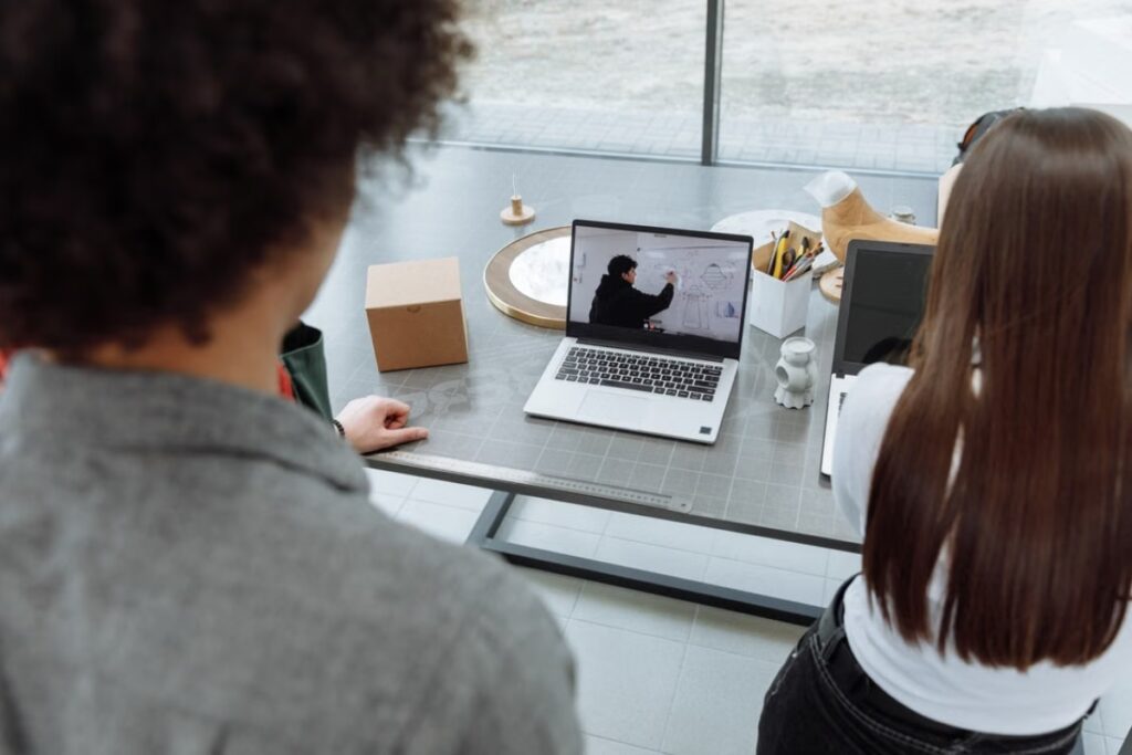 Vista superior traseira de duas pessoas, um homem de cabelos cacheados à esquerda e uma mulher de cabelos longos à direita, sentadas em uma mesa de trabalho compartilhada, assistindo a uma apresentação de vídeo em um notebook.