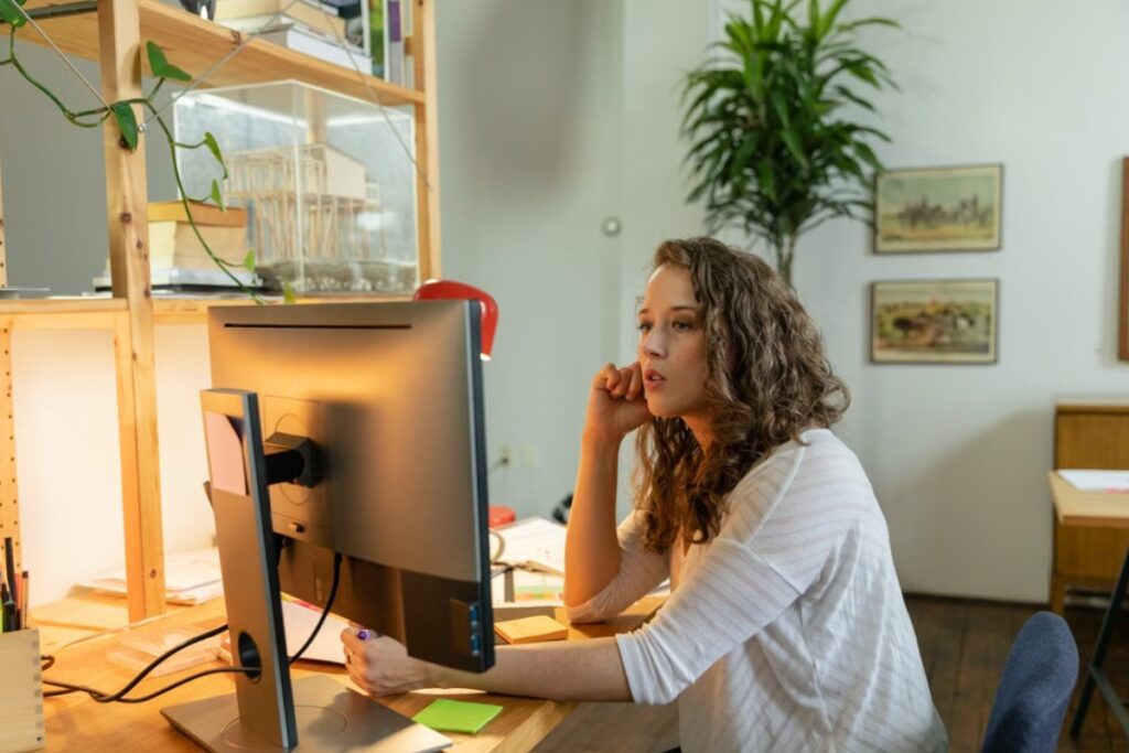 Mulher de cabelos cacheados e blusa branca sentada em frente a um monitor de computador em uma mesa de madeira, apoiando o queixo com uma das mãos sob iluminação interna suave.