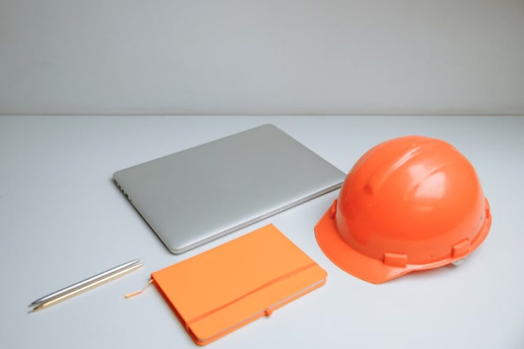 Notebook prata fechado, bloco de notas e canetas ao lado de um capacete de segurança laranja sobre uma mesa branca e limpa.