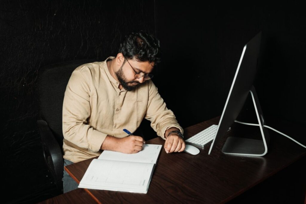 Homem barbudo de óculos e camisa bege sentado em uma mesa escura, focado enquanto escreve em um caderno aberto ao lado de um computador desktop Apple Mac sob fundo preto.