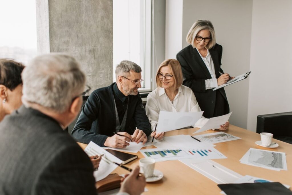 Um grupo de profissionais maduros reunido em torno de uma mesa de madeira, analisando gráficos corporativos e relatórios impressos, enquanto uma mulher em pé de blazer escuro faz anotações em uma prancheta.
