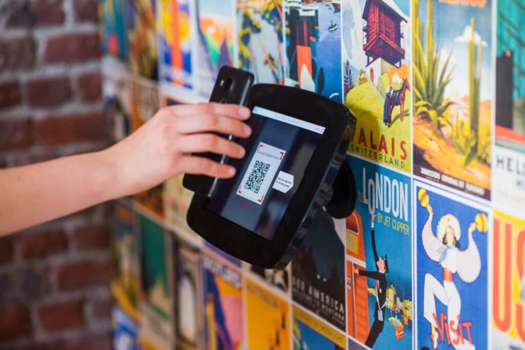 Fotografia em plano próximo focando na mão de uma pessoa segurando um smartphone preto para escanear um QR Code exibido em um dispositivo digital de leitura fixado em uma parede decorada com cartazes coloridos de viagens.