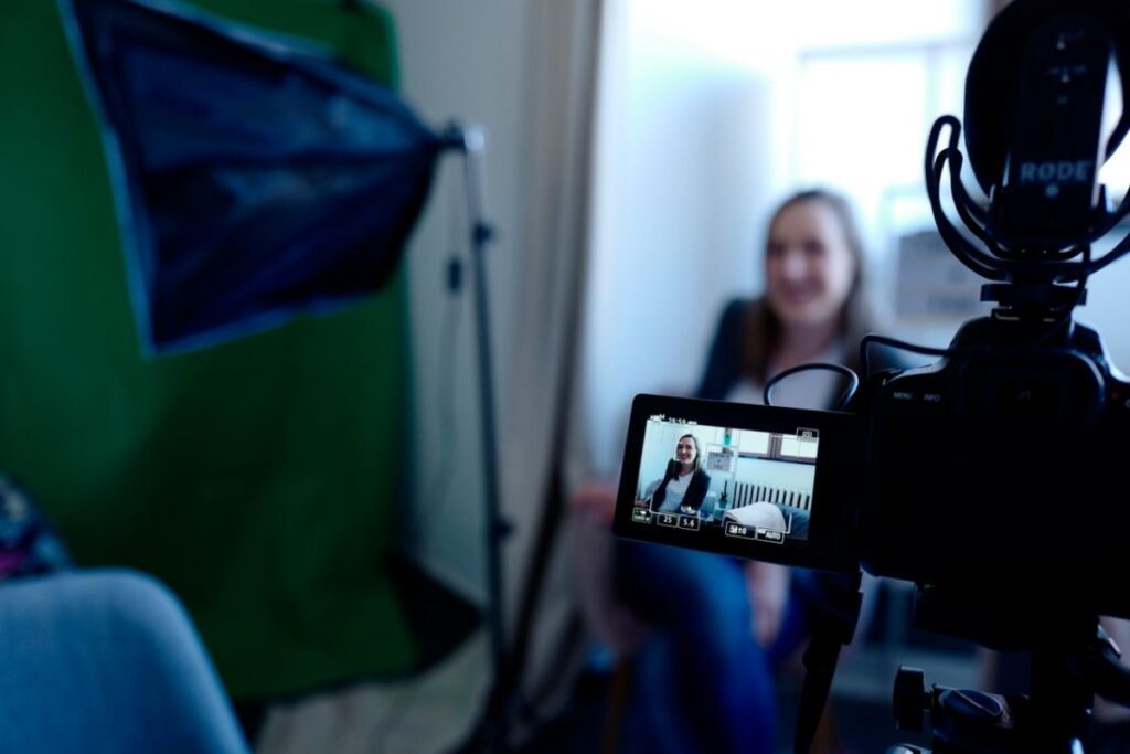 Close-up na tela acoplada de uma câmera DSLR filmando uma mulher sorridente ao fundo em um estúdio com fundo verde de chroma key e softbox de iluminação.