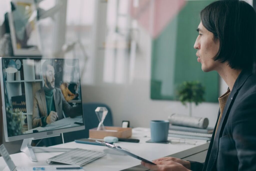 Vista lateral de um homem de terno conversando e olhando para a tela de um computador desktop que exibe outro homem de paletó em uma chamada de vídeo.