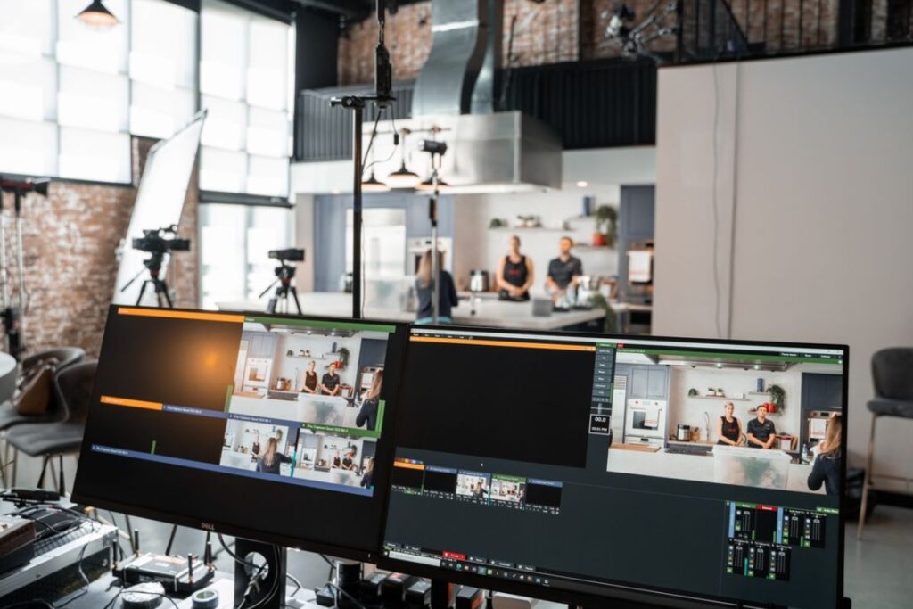 Dois monitores de computador exibindo um software de corte e transmissão ao vivo no primeiro plano, revelando ao fundo um estúdio com câmeras, iluminação e duas pessoas em uma bancada de cozinha.