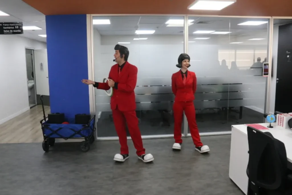 Dois apresentadores caracterizados em trajes vermelhos explicam a ferramenta 5W2H em um treinamento corporativo